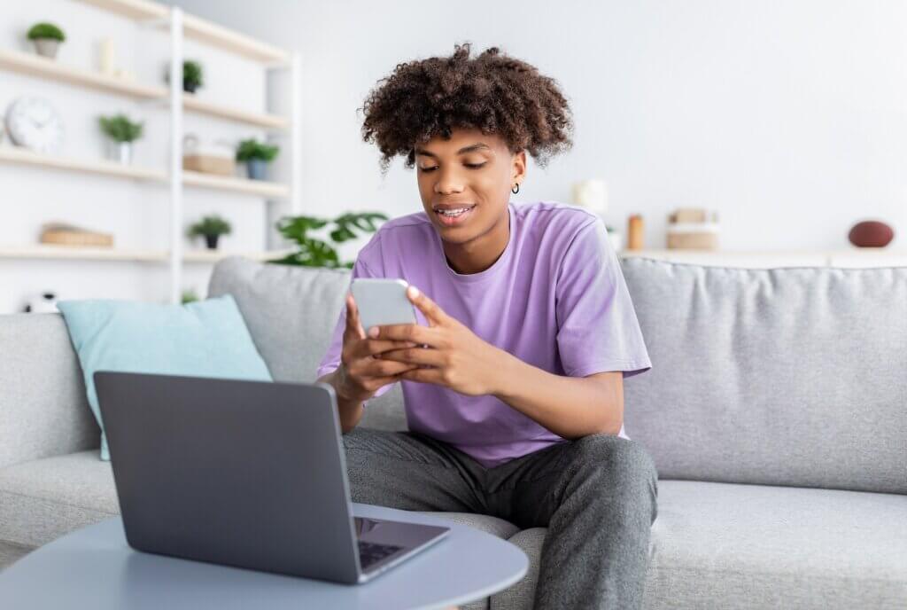 Happy young black teen in casual wear using smartphone and laptop, studying online from home